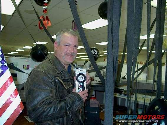 Man in leather jacket holds a ribbon award in an office decorated with balloons and streamers