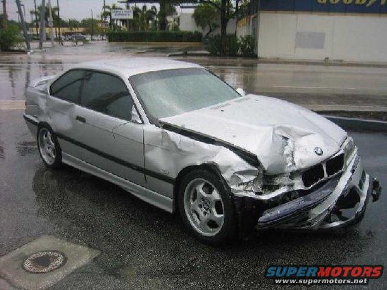 Silver 1995 BMW M3 coupe with crumpled front bumper and hood, parked on a wet street