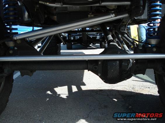 Undercarriage view of a modified off-road vehicle, showing suspension and drivetrain components. Another vehicle is visible in the background