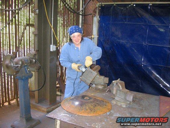 Woman in blue overalls and bandana using an angle grinder on a metal piece held in a vise