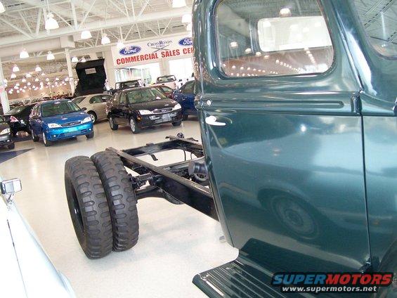 Close-up of a dark green truck chassis with dual rear wheels, other cars visible in background showroom