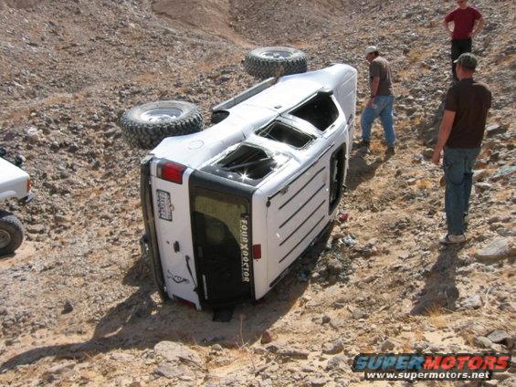 Overturned white SUV with large tires on a rocky desert trail. Two other vehicles and three people are visible