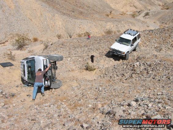 One SUV is on its side, being pulled by a rope attached to another SUV on a rocky hillside