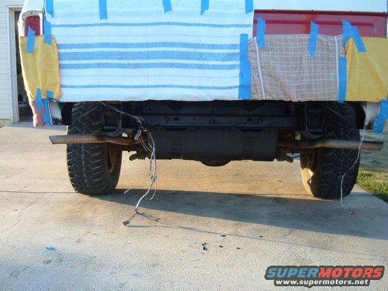 Rear axle, differential, and dual exhaust pipes of a truck, with wires hanging loose. The bed is covered with blue and white striped fabric and plaid fabric