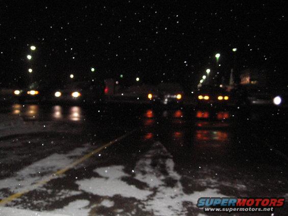 Multiple cars with headlights on, reflected on a wet road at night with falling snow