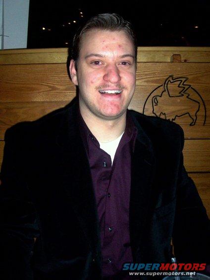 Man in a dark jacket and purple shirt smiles in front of a wooden wall with a buffalo logo
