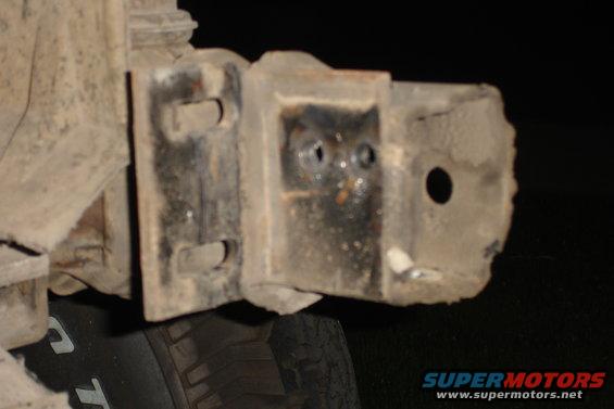 Close-up of a rusted, damaged metal bracket with two bolt holes and a larger circular hole, attached to a vehicle's undercarriage near a tire