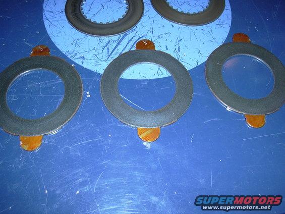 Several circular clutch plates with orange tabs on a blue surface. Supermotors logo in corner