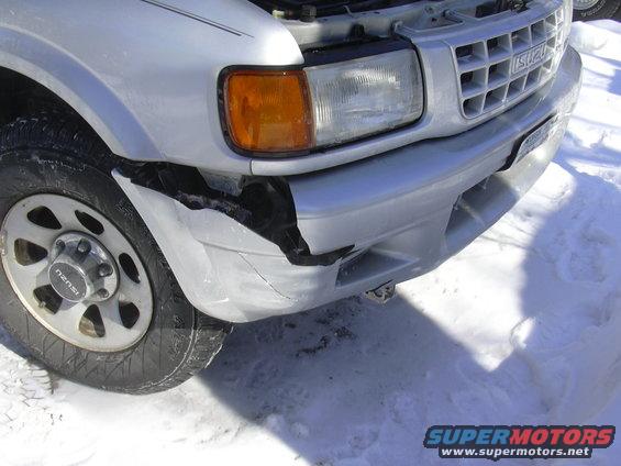 Front corner of a silver Isuzu Rodeo with a damaged bumper and fender. Snow on the ground