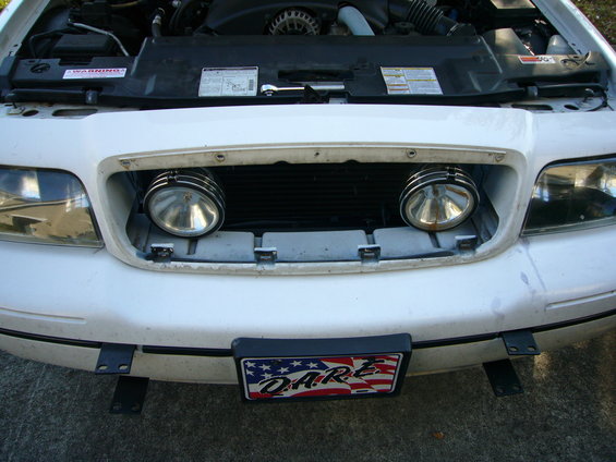 Front view of a white 2000 Ford Crown Victoria with the hood open, showing auxiliary lights installed in the grille area and a D.A.R.E. license plate frame