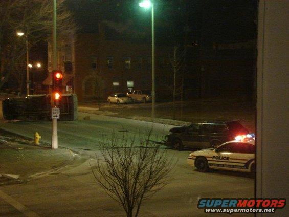 Overturned car and police cars at night, with traffic lights and buildings in background