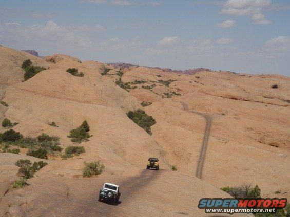 Two off-road vehicles on a desert road; one white, one yellow. Supermotors.net logo visible