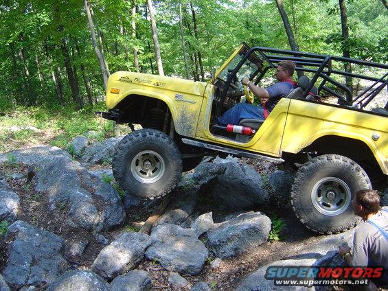 Yellow 1975 Ford Bronco driving over rocks on a wooded trail. Driver is visible