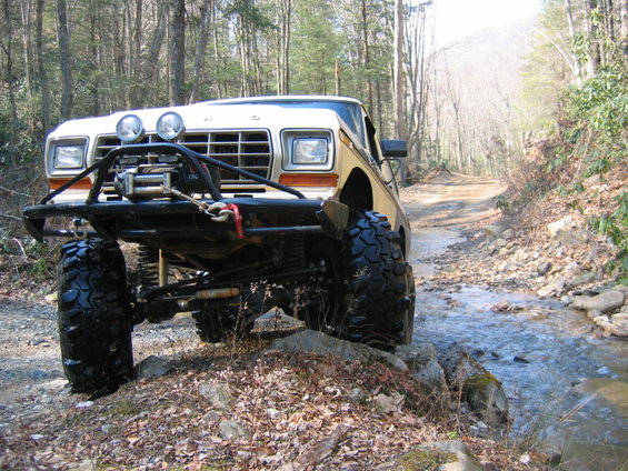 Modified 1978 Ford Bronco with large tires and winch on a dirt road crossing a stream