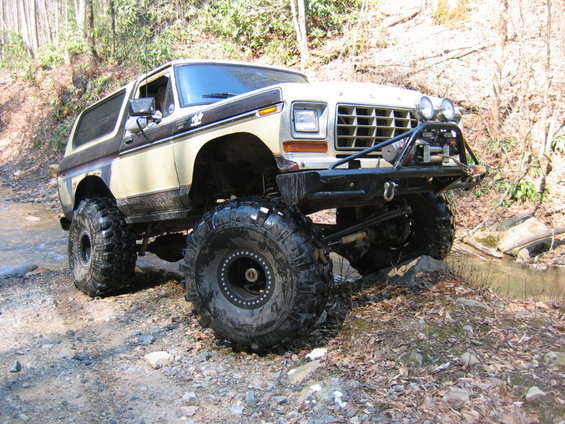 Lifted Ford Bronco on a dirt path with a stream nearby. It has large off-road tires and a custom bumper with lights and a winch