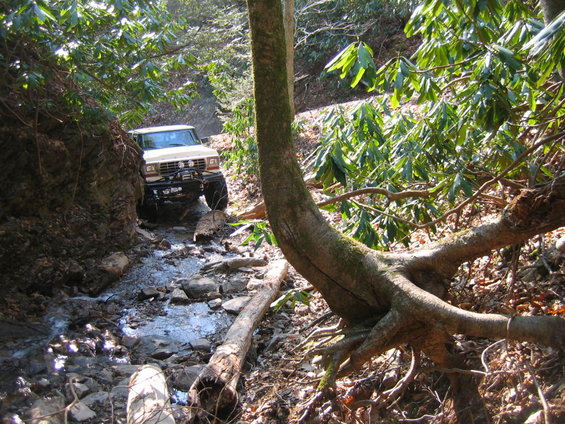 White off-road truck with winch driving through a rocky stream bed, front 3/4 view