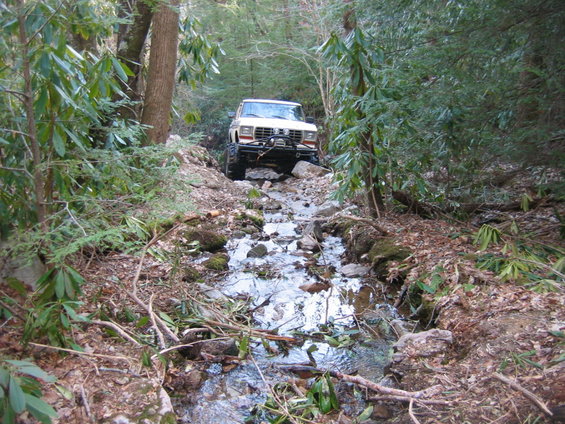 White off-road vehicle with winch, driving through a rocky stream bed in a forest