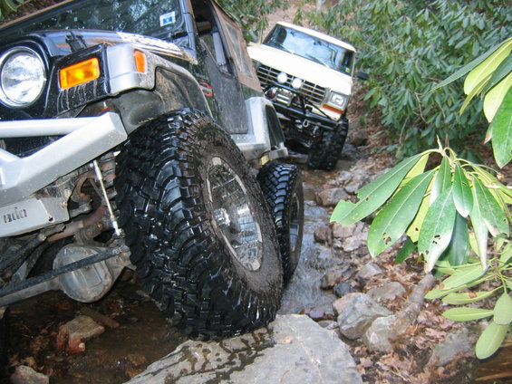 Black Jeep Wrangler and white Ford Bronco off-roading on a rocky, wooded trail with water