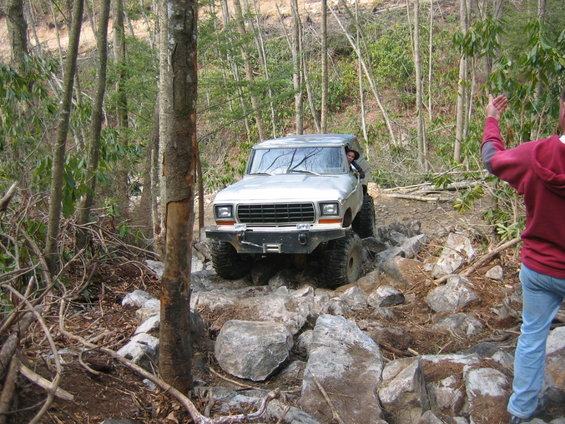 1978 Ford Bronco navigating a steep, rocky trail with trees on either side. Driver is visible