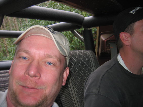 Man wearing a tan baseball cap smiles at the camera from the passenger seat of a vehicle, with another man in a black baseball cap visible in the background