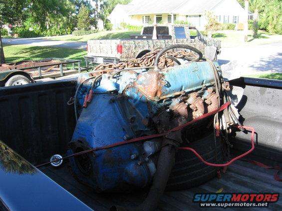 Blue engine with rusty parts and chains secured in a truck bed. A trailer is visible behind it