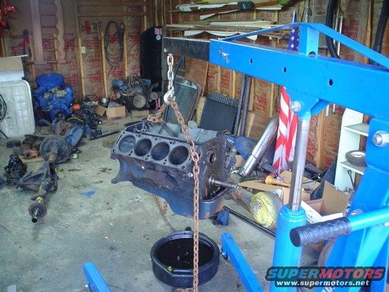 engine block hanging from an engine hoist in a garage with other parts around