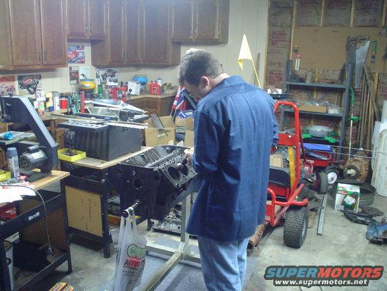 Person working on a black engine block mounted on a stand in a cluttered garage. A red cart is nearby