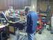 1976 Ford F-100 - Mechanic works on an engine block in a garage filled with tools and equipment.