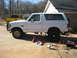 1996 Ford Bronco - A white 1996 Ford Bronco is parked on jack stands in a driveway, with a yellow pickup truck in the background.