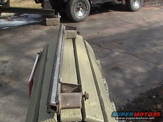 Close-up of a weathered wooden structure with metal brackets and bolts. A vehicle tire is visible in the background