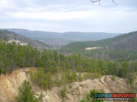 Mountainous terrain with pine trees and a dirt road winding through the valley under a cloudy sky. Watermark reads SUPERMOTORS