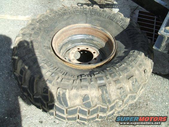 Off-road tire with aggressive tread pattern mounted on a rusty metal rim. Supermotors.net logo visible