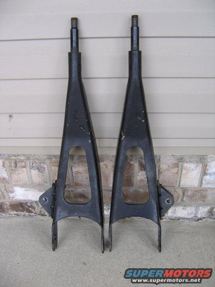 Pair of black control arms with triangular cutouts and mounting points, resting on concrete against a brick and siding wall
