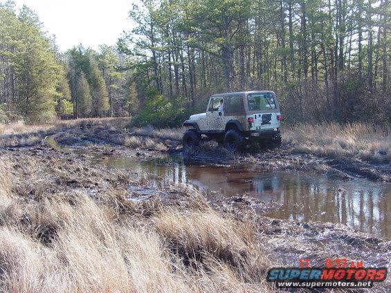 White Jeep Wrangler with a brown top driving through mud and water, trees in background