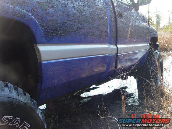 Blue pickup truck with silver trim, covered in mud, in shallow water. Side view