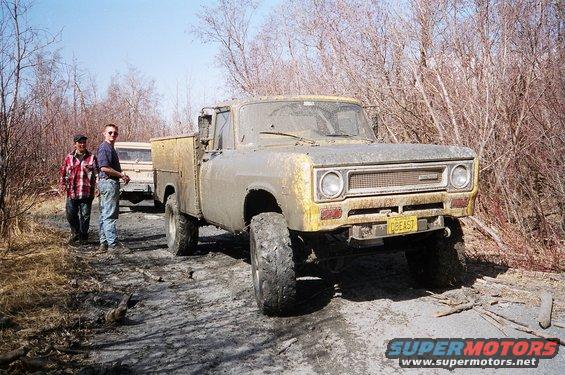 Yellow 1971 International pickup truck with a utility bed, covered in mud, parked on a dirt road with two men standing nearby