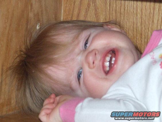 Baby with blonde hair and blue eyes smiling, wearing a white shirt with pink trim