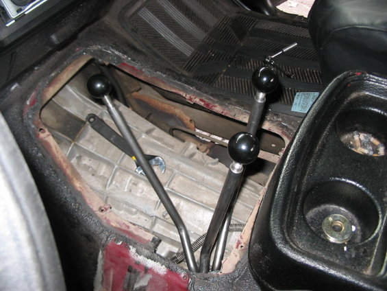 Vehicle interior showing a transmission tunnel with three manual shifters and a wrench. Red paint is visible under the black trim