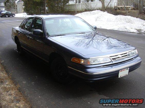 Dark blue sedan parked on asphalt with snow in background. Another car is visible in the distance