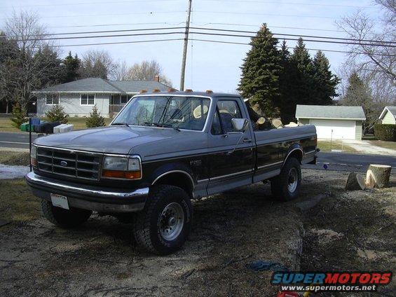 Gray and black Ford F-Series pickup truck with logs in the bed, parked on dirt