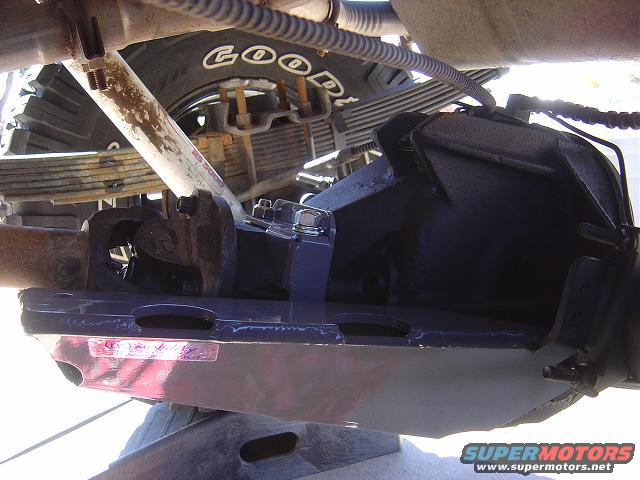 Underside view of a vehicle's rear axle, driveshaft, leaf springs, and shock absorber. A 'GOOD YEAR' tire is visible