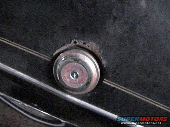 Close-up of a circular car emblem with red and silver accents, mounted on a dark surface with some rust around the edges