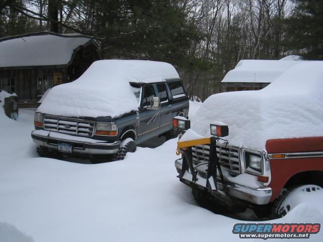 Dark blue SUV with snow plow attachment in front, parked next to a red vehicle covered in snow