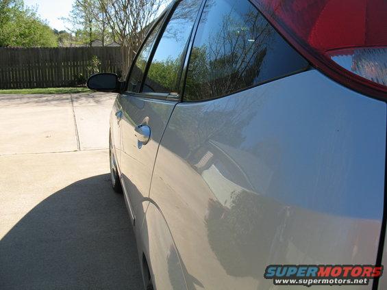 Silver car side profile, showing door handle and rear window. Trees reflected in glass