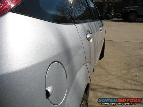 Side view of a silver sedan's rear door and fuel cap. Another SUV is parked in the background