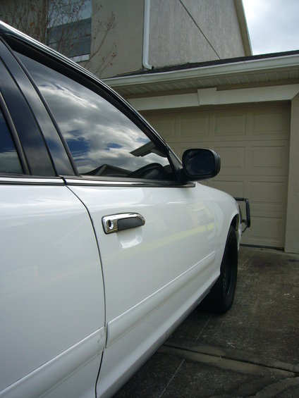 Side view of a white sedan parked in front of a garage. Shows door handle and side mirror