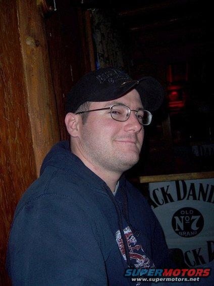 Man in glasses and a dark blue hoodie with a black baseball cap, seated indoors with a Jack Daniel's sign in the background