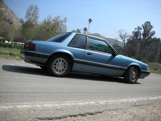 Blue 1990 Ford Mustang coupe parked on the side of a road with hills and trees in the background