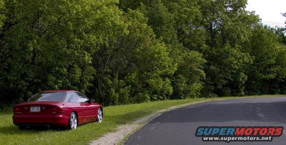 Red 1993 Ford Probe rear view, parked on grassy shoulder next to a winding road