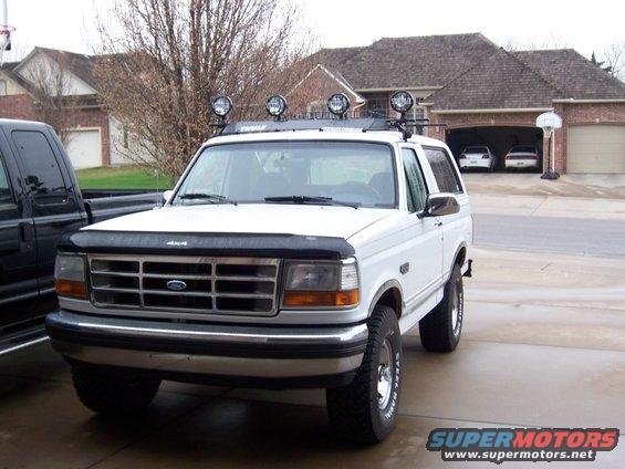 White Ford Bronco, front view, with roof rack and four spotlights. A black truck is partially visible to the left
